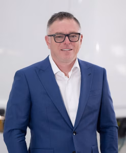 Professional middle-aged man wearing black glasses, blue suit jacket, and white shirt smiling against a plain background