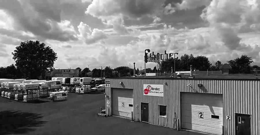Black and white image of an industrial warehouse exterior with multiple loading bays and trucks parked outside under a cloudy sky