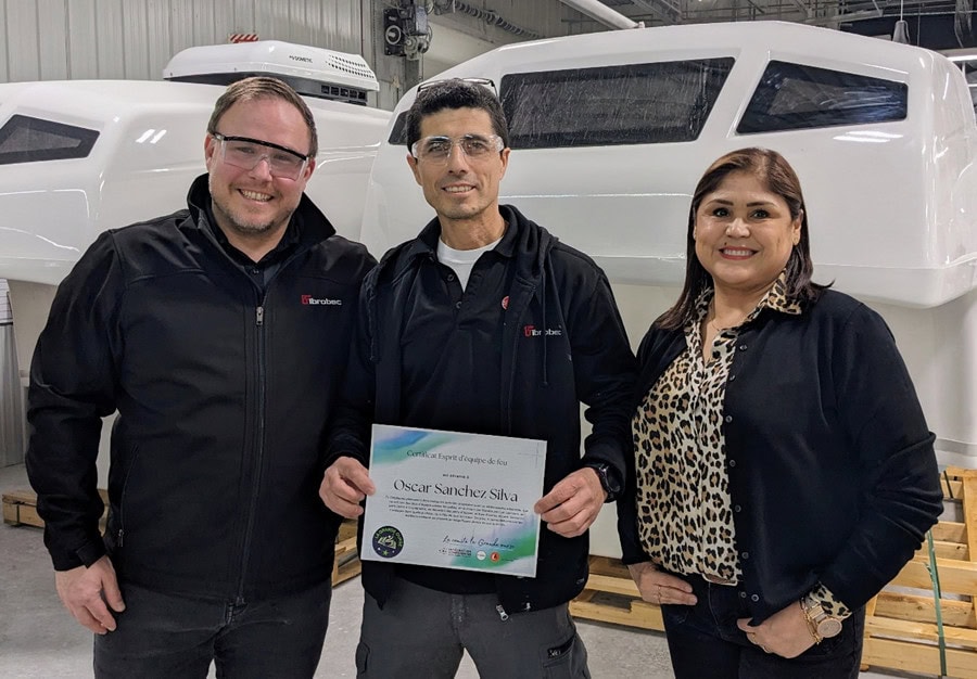 Three people smiling indoors, one holding a certificate with the name Oscar Sanchez Silva, standing in front of large white industrial equipment.
