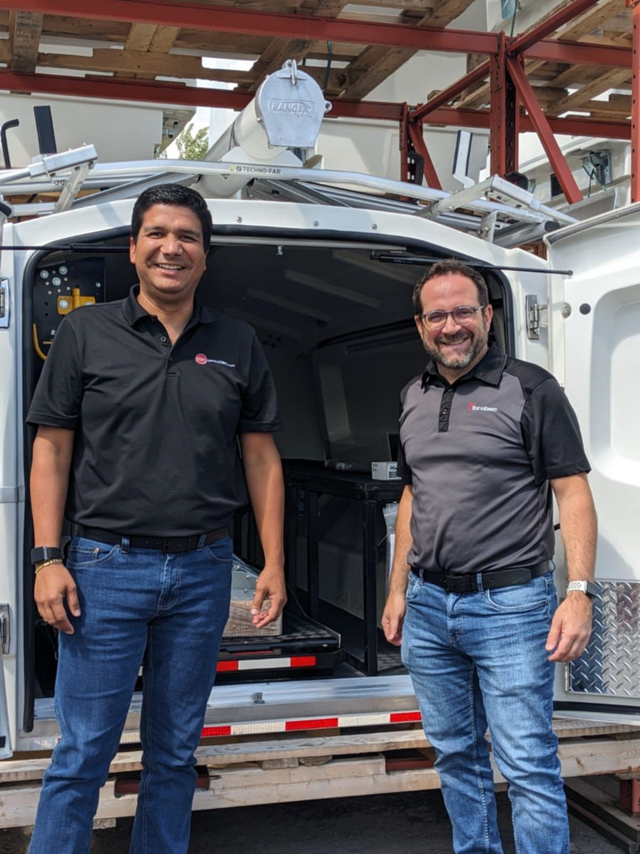 Two smiling men standing in front of an open service vehicle, wearing casual business attire