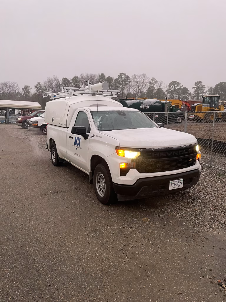 White Chevrolet utility truck with JCI company logo parked on a gravel lot near construction equipment with headlights on on an overcast day