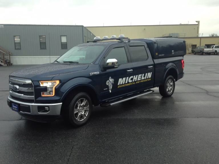 Dark blue Ford F-150 pickup truck with Michelin Environmental and Prevention logo parked on wet pavement in front of industrial buildings