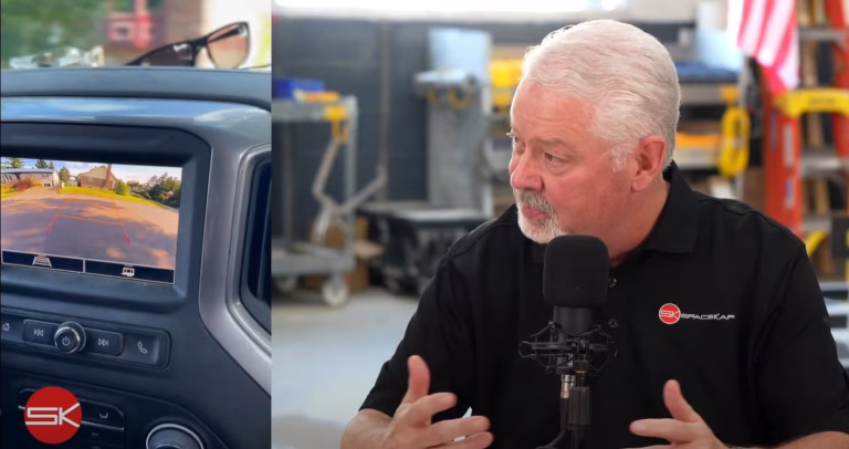 Man with white hair and beard speaking into a microphone in a garage or workshop setting with a backup camera display on a vehicle dashboard