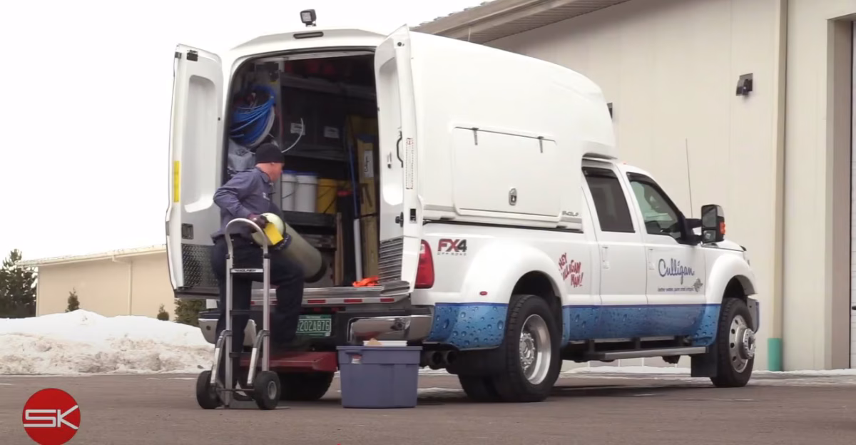 Technician unloading equipment from a white Culligan service truck parked outside a building on a snowy day