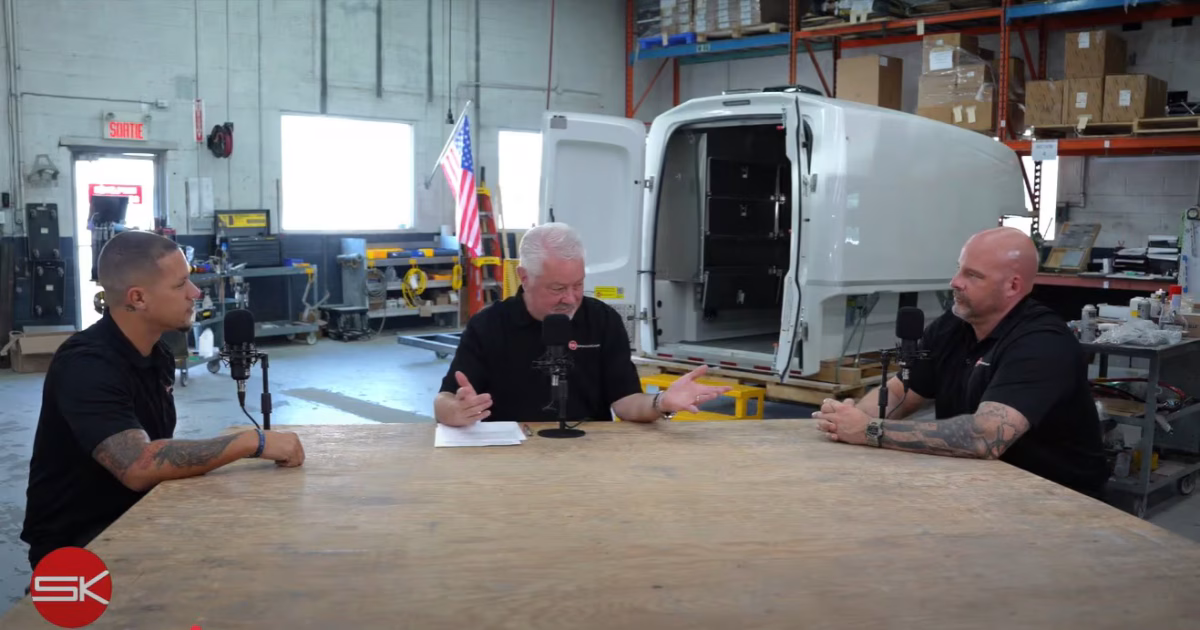 Three men having a discussion at a table in a workshop with a white truck cargo box and American flag in the background