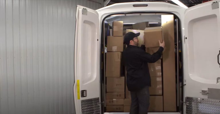 Man loading cardboard boxes into a white delivery van with rear doors open inside a warehouse