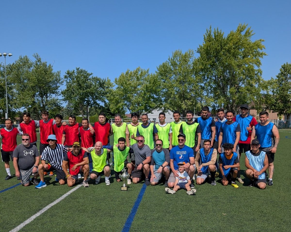 Group photo of soccer players in red, yellow, and blue jerseys posing on a green field with trees in the background under a clear blue sky
