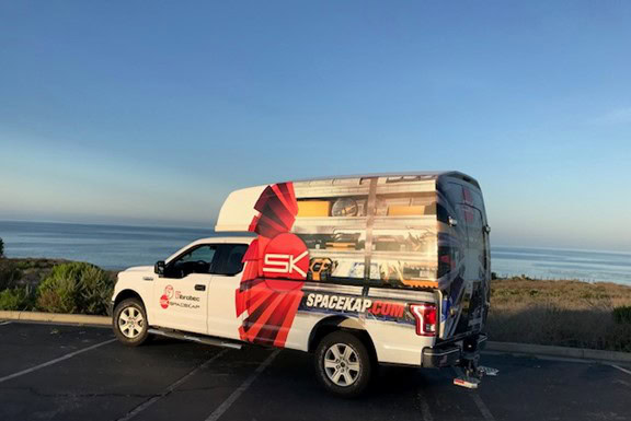SpaceKap branded white pickup truck with red and black logo parked by the ocean during sunset