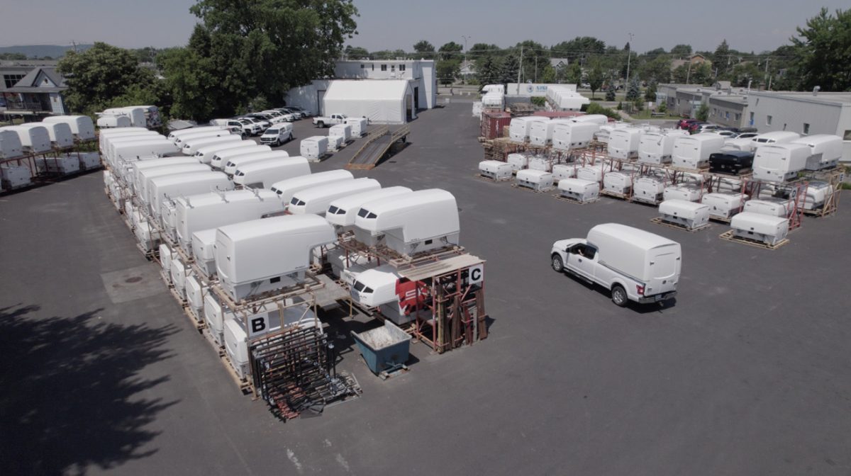 Aerial view of a large outdoor storage area with multiple white truck SpaceKaps organized and stacked on metal racks, and a white pickup truck driving through the paved lot.