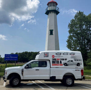 White pickup truck with a large white service van cap parked near a lighthouse and a Pure Michigan welcome sign