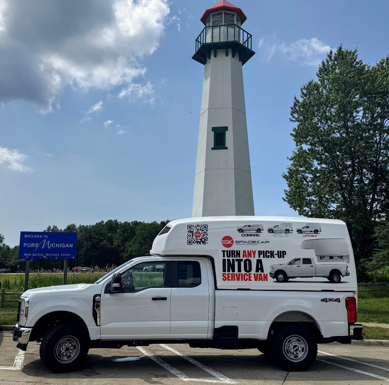 White pickup truck with a large white service van cap parked near a lighthouse and a Pure Michigan welcome sign