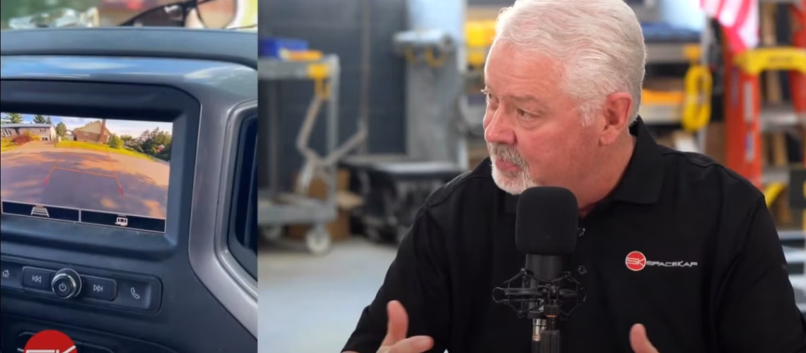 Man with white hair and beard speaking into a microphone in a garage or workshop setting with a backup camera display on a vehicle dashboard