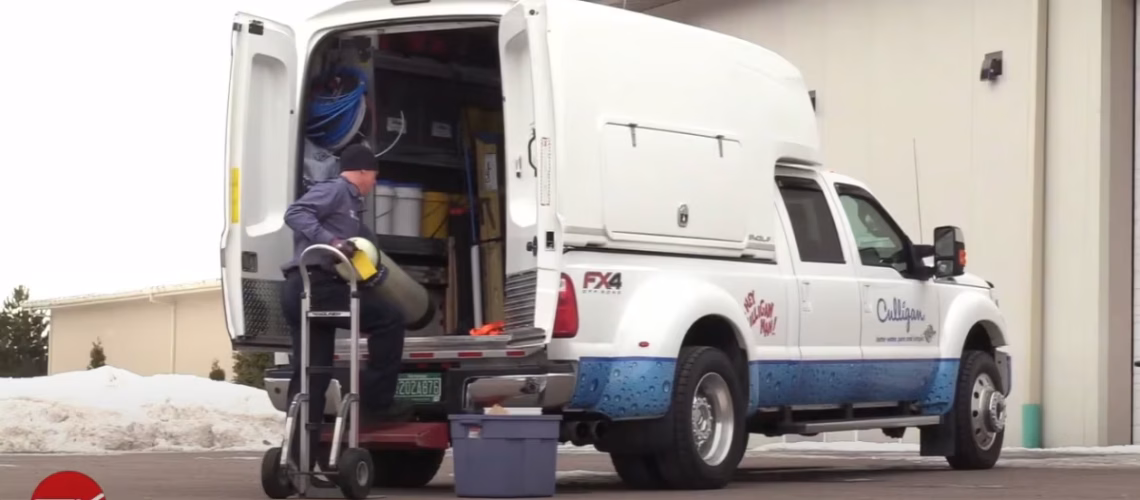 Technician unloading equipment from a white Culligan service truck parked outside a building on a snowy day