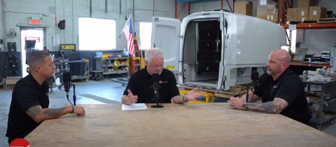 Three men having a discussion at a table in a workshop with a white truck cargo box and American flag in the background