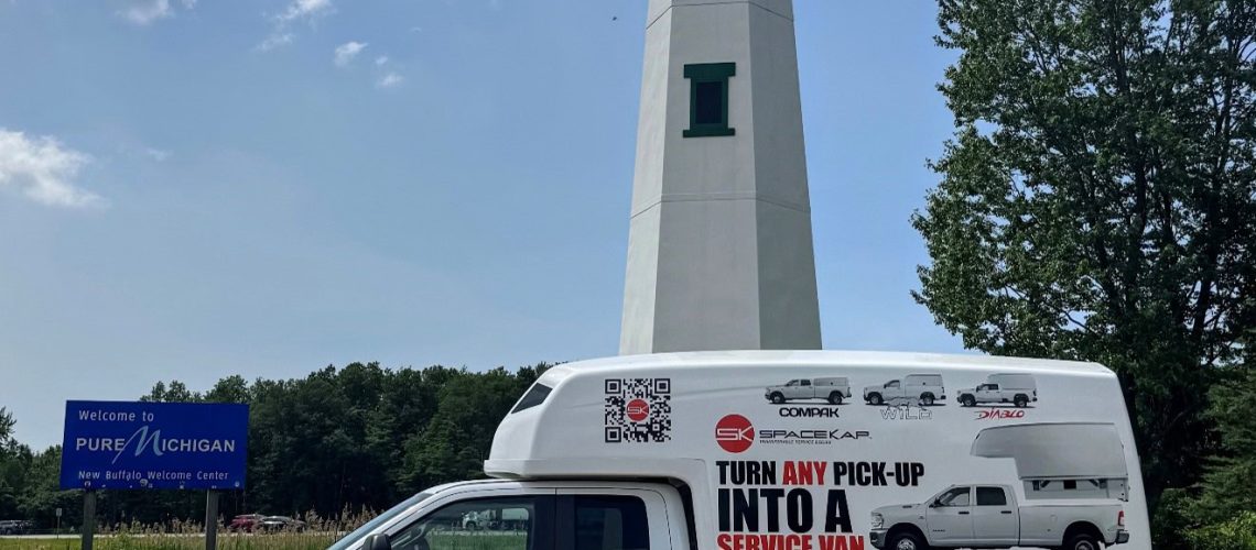 White pickup truck with a large white service van cap parked near a lighthouse and a Pure Michigan welcome sign