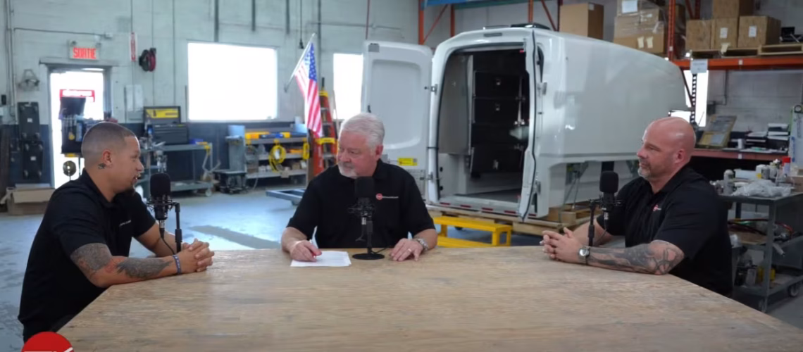 Three men in black shirts sitting around a wooden table with microphones in a workshop with an open white vehicle canopy in the background