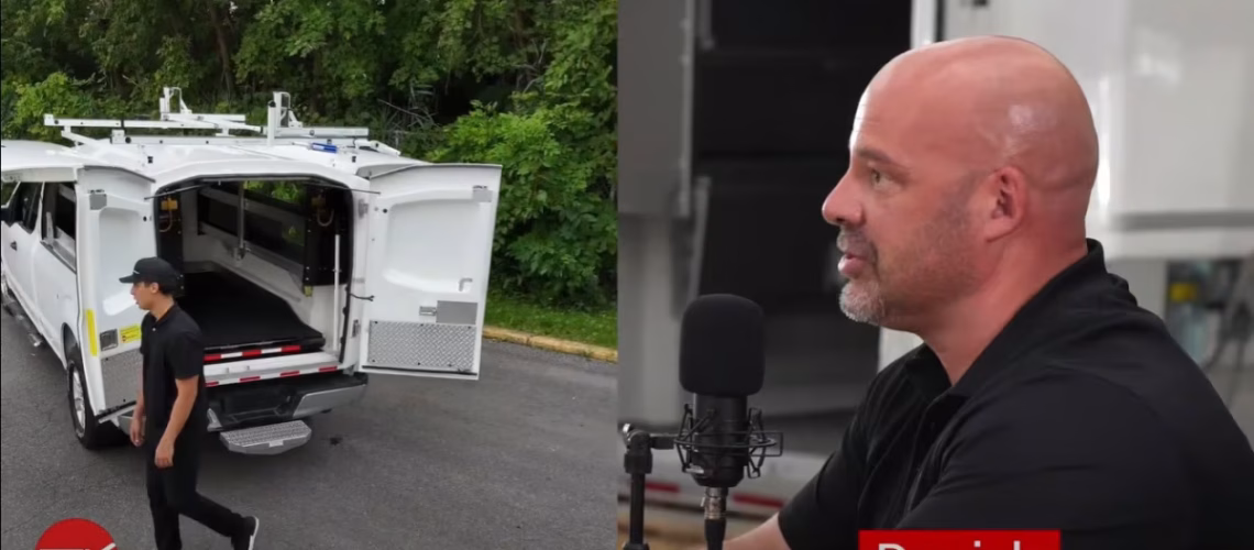 Man speaking into microphone during interview alongside white utility truck with open rear doors